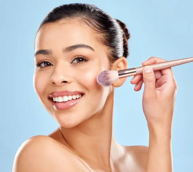 Une femme souriante utilise un pinceau pour appliquer du correcteur maquillage sur sa joue.