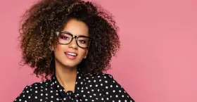 Une jeune femme aux cheveux bouclés porte un maquillage éclatant et des lunettes noires, souriant sur fond rose.