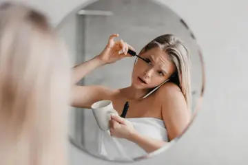 Une femme, enroulée dans une serviette, applique son mascara tout en tenant une tasse et en parlant au téléphone. Le mascara qui coule sur son visage témoigne de sa matinée mouvementée.