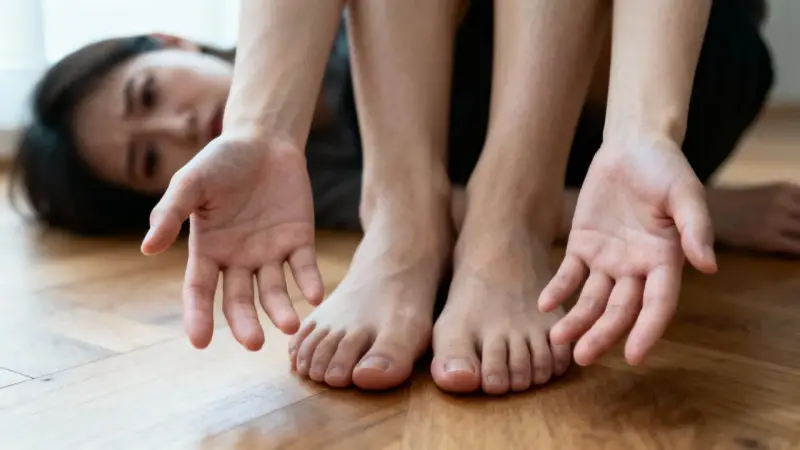 Femme aux mains tendues et pieds nus sur un parquet. Ses ongles semblent fragiles, peut-être un signe de carence en vitamine B12.