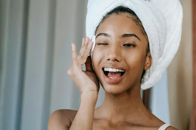 Une jeune femme avec une serviette sur la tête, appliquant un soin sur son visage. Elle semble heureuse, peut-être grâce aux bienfaits du collagène ou de l'acide hyaluronique.